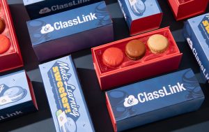 Branded ClassLink macaron gift boxes in blue and red packaging arranged on a dark surface, with one open box displaying three assorted macarons.
