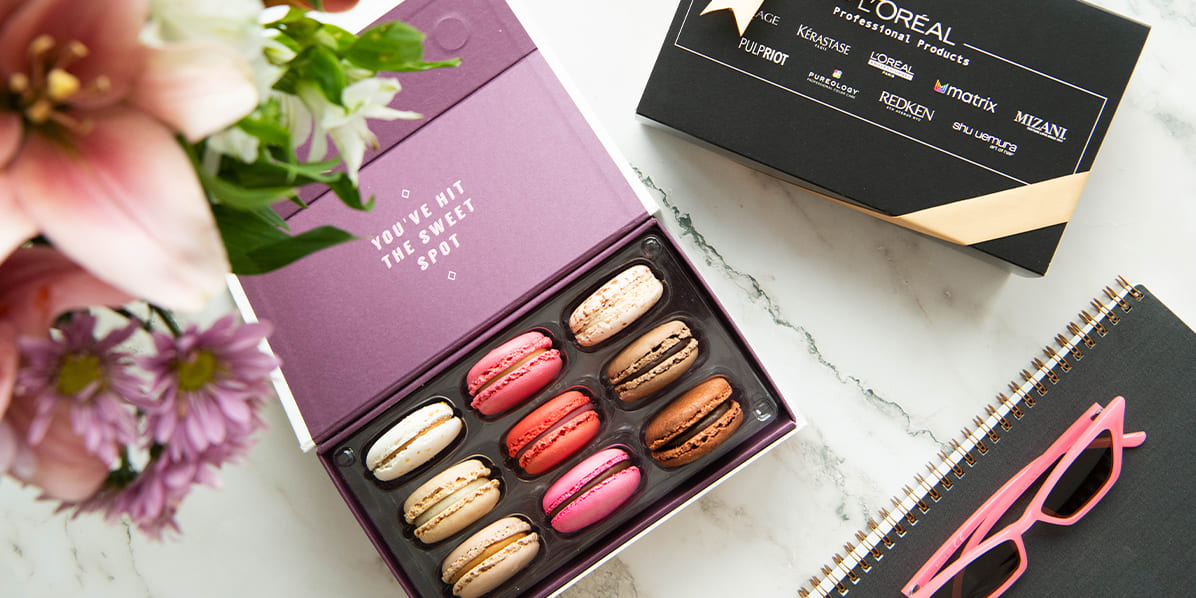 Open purple macaron gift box with assorted flavors on a marble surface beside a black L’Oréal Professional Products branded box with gold ribbon, flowers, a notebook, and pink sunglasses.