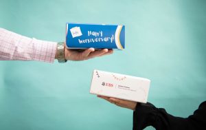 Two hands holding branded gift boxes against a teal background, one reading “Happy Workiversary!” and the other displaying a corporate logo and birthday-themed design.