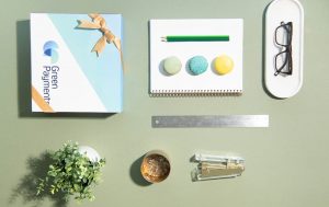 Flat lay of a branded gift box alongside office essentials, including a notebook with macarons, pencils, glasses case, ruler, plant, and desk accessories on a soft green background.