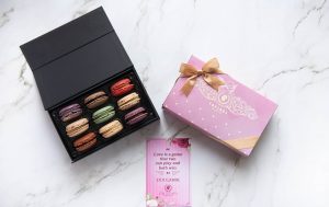 Open black box of assorted macarons beside a pink ribbon-tied gift box and a romantic quote card on a marble background.