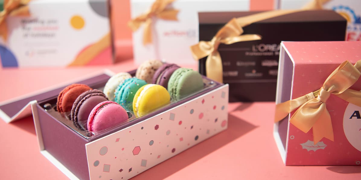 Open gift box of colorful macarons on a pink background, surrounded by branded ribbon-tied gift boxes.