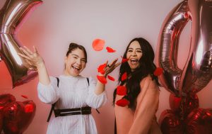 Two women laughing and tossing red rose petals, surrounded by heart-shaped balloons in a festive Valentine’s Day setting.