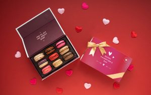 Open heart-themed macaron gift box with assorted macarons beside a Valentine’s Day card on a red background with scattered heart decorations.