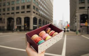 Hand holding an open box of assorted colorful macarons on a city street with buildings in the background.