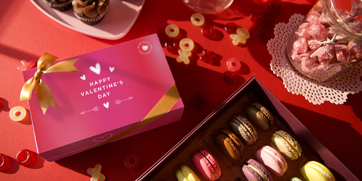 Valentine’s Day macaron gift boxes and desserts styled on a red background with heart decorations and candies.