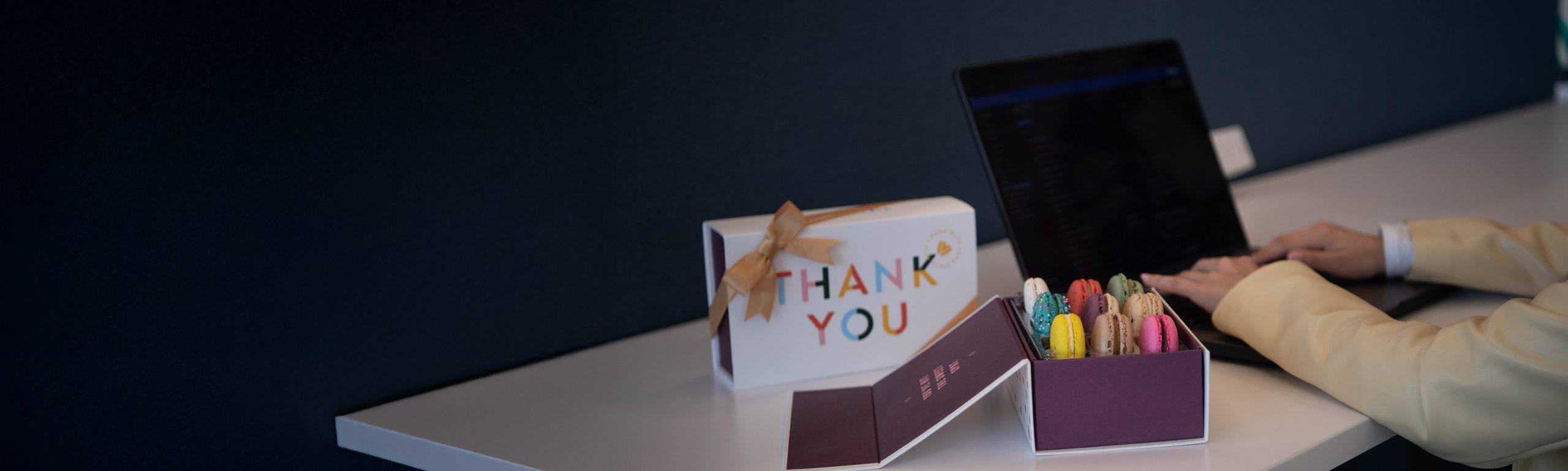 Woops! New Year’s macaron gift boxes with gold ribbons displayed on a shiny surface surrounded by disco balls, champagne glasses, and festive decorations.