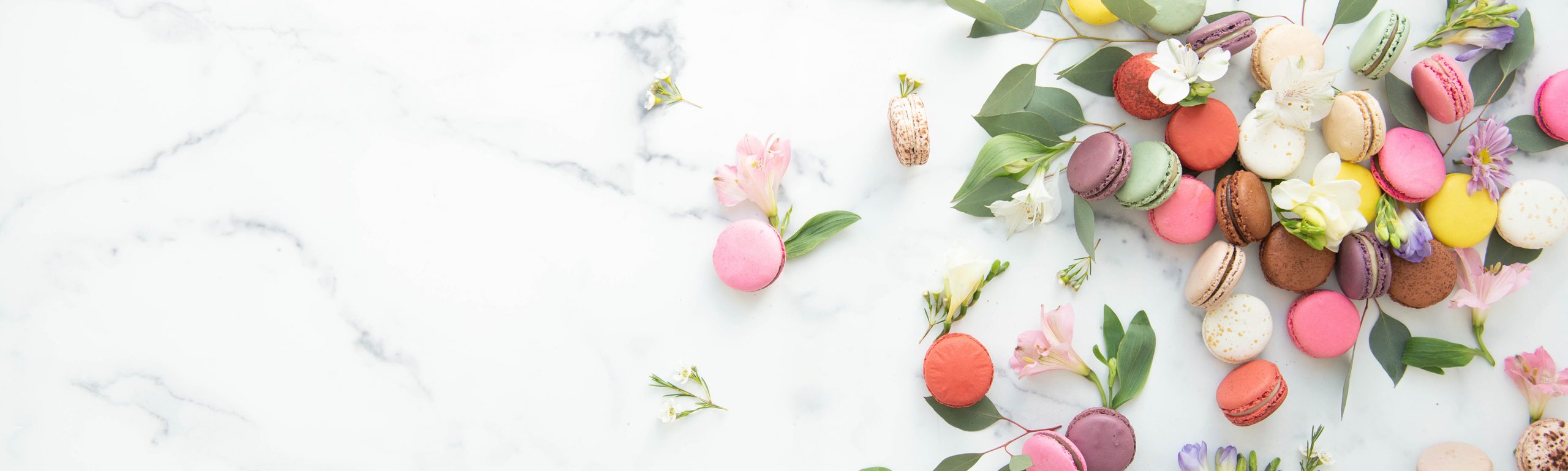 Assorted pastel macarons styled with fresh flowers and green leaves on a light marble surface.