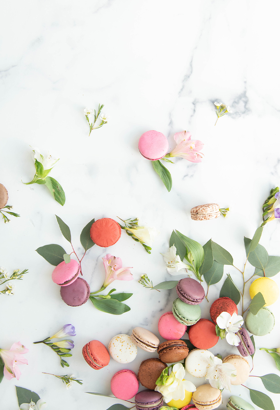 Assorted pastel macarons styled with fresh flowers and green leaves on a light marble surface.